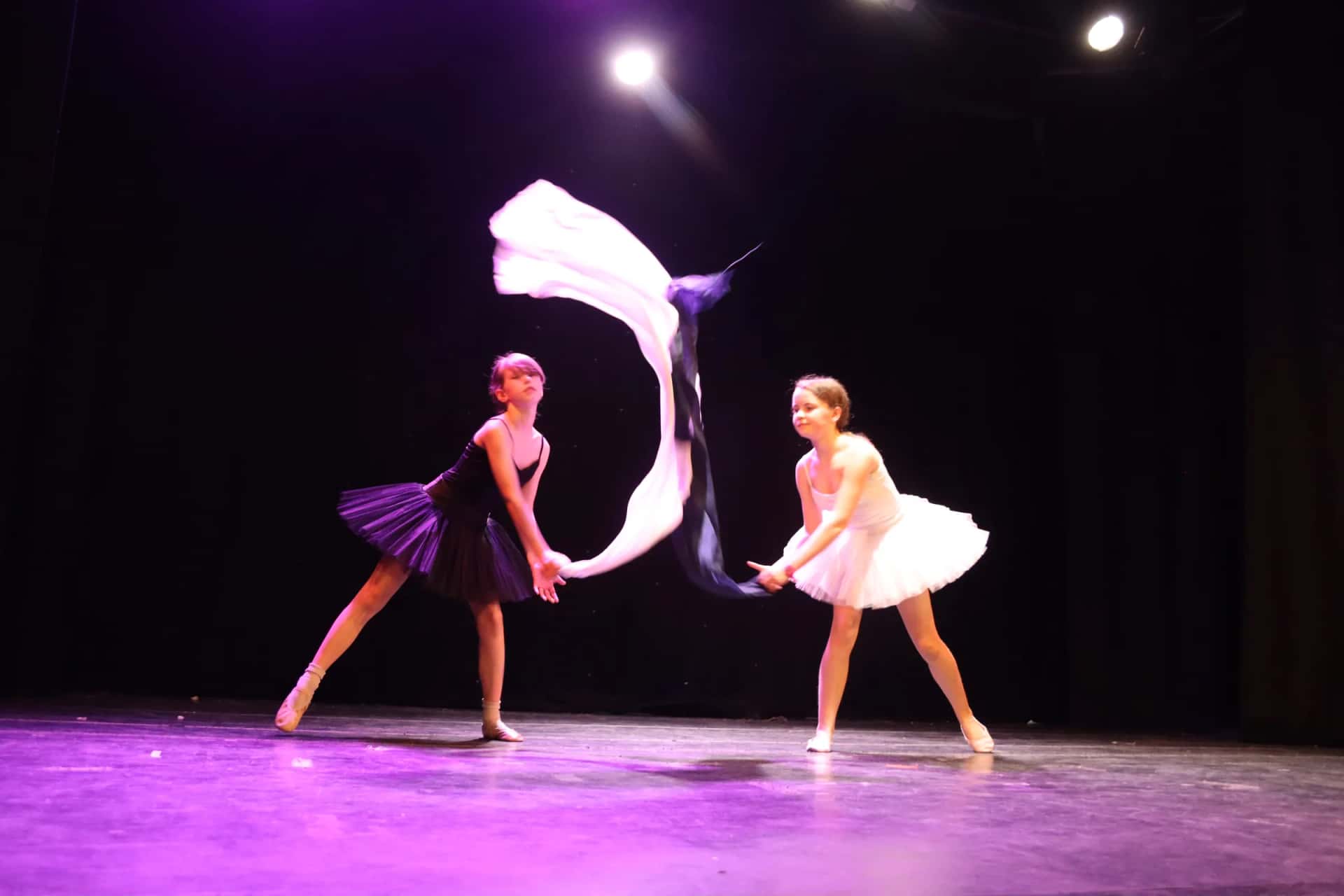 Two young ballet dancers performing a duet on stage, one swirling a white silk under purple stage lights